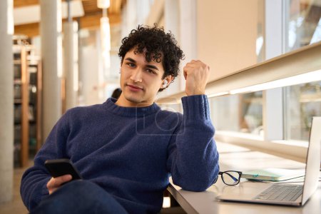 Photo for Handsome positive young Hispanic man, looking at camera, sitting at desk with laptop and holding a mobile phone in his hands. Male student in the library campus. People. Education. Learning concept - Royalty Free Image