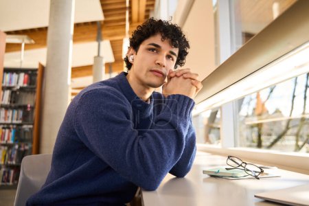 Photo for Thoughtful young Latin American male student, wearing earphones, listens online lecture, sitting at desk with laptop and studying in the modern innovation university library campus. Education concept - Royalty Free Image