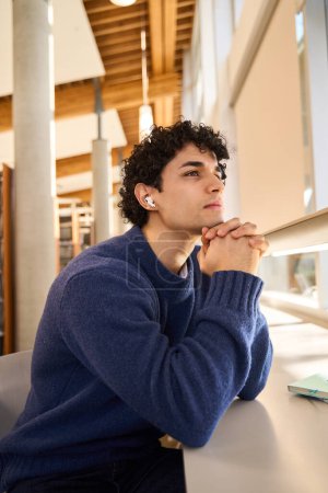 Photo for Pensive young dark-haired multi-ethnic male student, wearing earphones, listens online lecture, in the modern university library campus. Education. Erudition. Online studying and people concept - Royalty Free Image