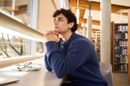 Photo for Confident portrait of a smart Hispanic male student sitting at desk in a modern library campus, attentively listening to a broadcasted online lesson on earphones, in the university library campus - Royalty Free Image