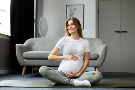 Téléchargez les photos : Portrait d'une femme enceinte souriante assise sur le sol et touchant son ventre. Concept de yoga de grossesse - en image libre de droit