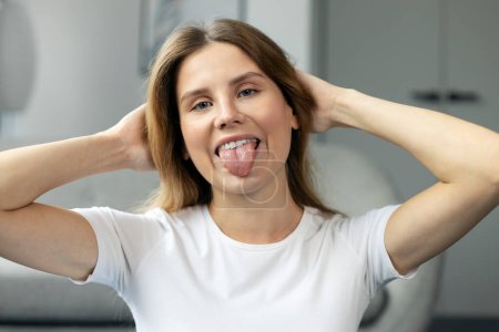 Foto de Retrato de una hermosa joven hembra mirando a la cámara y mostrando la lengua, posando contra la sala de estar. Concepto de feminidad y belleza - Imagen libre de derechos