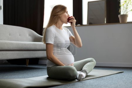 Foto de Mujer embarazada joven comiendo manzana roja practicando yoga sentada en pose de loto en casa. Yoga del embarazo, dieta, concepto de comida saludable - Imagen libre de derechos