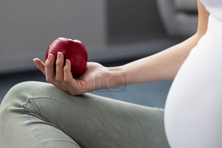 Foto de Mujer embarazada practicando yoga sentada en el suelo y sosteniendo manzana roja en casa, enfoque selectivo. Embarazo, estilo de vida saludable, concepto de dieta - Imagen libre de derechos