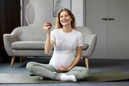 Photo for Young smiling pregnant woman practicing yoga sitting in lotus pose, holding red apple at home. Pregnancy yoga, healthy food concept - Royalty Free Image
