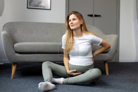 Photo for Young pregnant woman sitting on floor and touching her belly, feeling neck pain. Pregnancy concept - Royalty Free Image