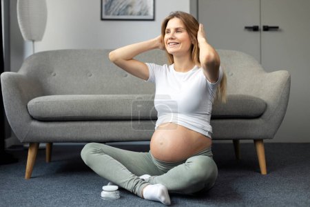 Photo for Smiling pregnant woman doing exercises, meditation, sitting in lotus position looking away at home. Pregnancy yoga concept - Royalty Free Image