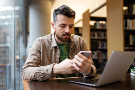 Photo for Handsome bearded student sitting at desk, using smartphone, shopping online, watching video or checking message. Attractive man sitting at desk with laptop. Concept of online learning - Royalty Free Image
