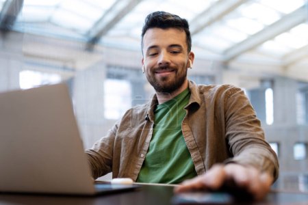 Téléchargez les photos : Un homme d'affaires souriant et séduisant utilise un ordinateur portable, un ordinateur sur son lieu de travail. Le mec d'âge moyen dans les écouteurs écoute des conférences en ligne, discute en ligne, écoute de la musique, les médias sociaux - en image libre de droit