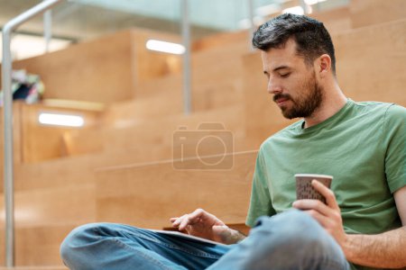 Téléchargez les photos : Portrait d'un homme hispanique de taille moyenne utilisant une tablette numérique en regardant des vidéos assis sur les marches du campus universitaire - en image libre de droit