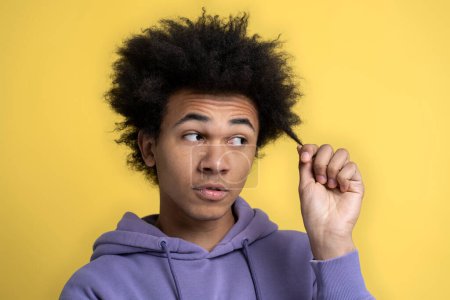 Téléchargez les photos : Triste bouclé homme afro-américain tenant les cheveux bouclés abîmés, regardant loin isolé sur fond jaune, studio tourné. Concept de soins capillaires - en image libre de droit
