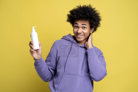 Foto de Sonriente atractivo hombre afroamericano sosteniendo una botella blanca de champú hidratante nutritivo contra la caspa sobre un fondo amarillo aislado. Cuidado del cabello y tratamiento médico de la caída del cabello - Imagen libre de derechos