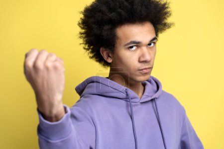 Foto de Hombre afroamericano enojado sosteniendo el puño apretado listo para luchar aislado sobre fondo amarillo. Estudiante agresivo preparándose para atacar, disparo de estudio - Imagen libre de derechos