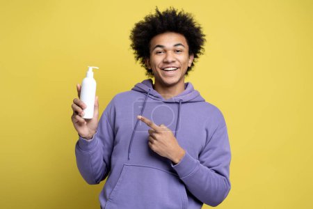 Foto de Joven hombre afroamericano sonriente sosteniendo la botella de champú para el cabello, señalando con el dedo, mirando a la cámara aislada sobre fondo amarillo, filmado en estudio. Cuidado del cabello, concepto de rutina matutina - Imagen libre de derechos