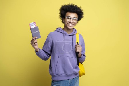 Foto de Attractive smiling African American man holding passport with boarding pass ticket isolated on yellow background. Happy tourist ready for flight. Vacation, travel concept - Imagen libre de derechos
