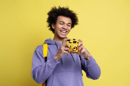 Photo for Happy curly haired guy photographer, journalist or traveler with yellow backpack, holding digital camera and smiling looking at camera, before taking photos isolated on yellow background - Royalty Free Image