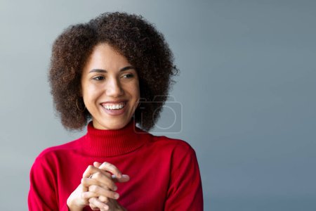 Foto de Retrato de cerca de una joven afroamericana alegre vestida con suéter rojo, sonriendo con cuidado, mirando hacia otro lado, de pie contra una pared gris con espacio libre para anuncios, expresando emociones positivas. - Imagen libre de derechos