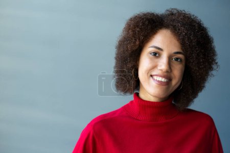 Close-up portrait of a confident multi-ethnic woman with stylish afro hairstyle, wearing red casual sweater, smiling cutely looking at camera, standing against a gray wall background. People. Emotions