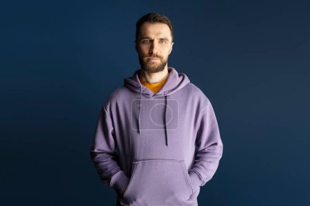 Photo for Confident portrait of a handsome middle-aged Caucasian bearded man, wearing purple hoodie, holding hands in pockets, looking confidently at camera over isolated dark blue background. People concept - Royalty Free Image