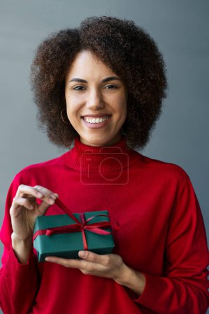 Téléchargez les photos : Belle jeune femme afro-américaine portant un pull rouge décontracté, souriant en regardant la caméra, tenant une jolie boîte cadeau enveloppée dans du papier d'emballage noir avec nœud noué rouge, sur fond de mur gris - en image libre de droit