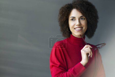 Foto de Mujer joven y elegante, señora de negocios confiada, gerente ejecutivo, empresario freelancer sonríe mirando pensativamente a un lado, sosteniendo elegantes gafas enmarcadas rojas en sus manos sobre el fondo de la pared gris - Imagen libre de derechos
