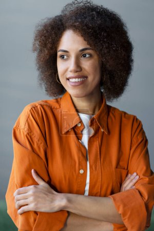 Foto de Elegante y pensativa mujer multiétnica de piel oscura con cabello afro, vestida con una camisa casual naranja, sonríe, mirando hacia un lado, de pie con los brazos cruzados sobre un fondo gris. Primer plano - Imagen libre de derechos