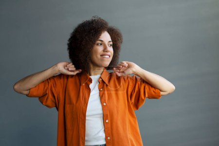 Photo for Portrait of a relaxed pensive African American young woman, dressed in casual orange shirt, dreamily looking aside, standing against a gray wall background at daylight. People. Daydreaming. Lifestyles - Royalty Free Image