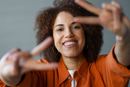 Photo for Headshot of positive smiling African American curly haired young woman smiling with a beautiful toothy smile, looking at camera, gesturing, showing peace signs with her fingers. People. Body language - Royalty Free Image
