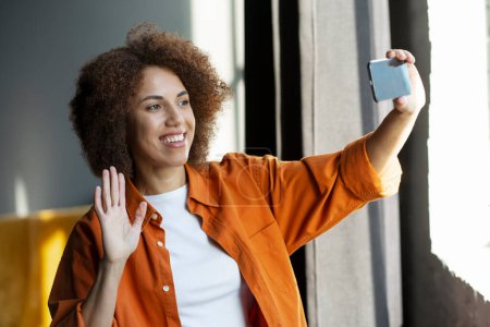 Photo for Pleasant young ethnic woman holding smartphone in her outstretched hand, greeting her interlocutor, waving hello, enjoying online meeting or conference by video link. Communication across the distance - Royalty Free Image