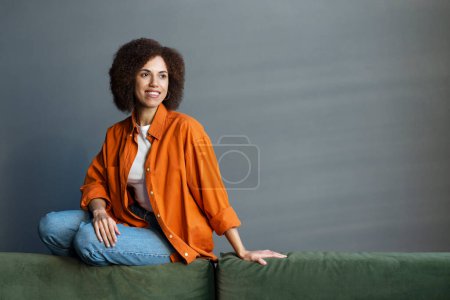 Foto de Retrato de una encantadora mujer afroamericana feliz pensativa, mirando hacia otro lado, sentada en un sofá en la sala de estar con una hermosa sonrisa y expresiones de la cara feliz. Gente. Estilos de vida. Ocupaciones - Imagen libre de derechos