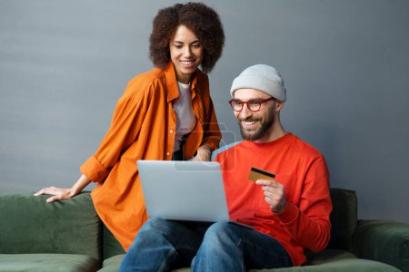 Schöner kaukasischer bärtiger Mann in lässiger Kleidung und Brille, der mit seiner Freundin auf einem Sofa sitzt, Kreditkarte in der Hand, Laptop benutzt, per Internet-Banking bezahlt, online einkauft