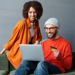 Handsome Caucasian bearded man in casual clothes and wearing glasses, sitting on a sofa with his girlfriend, holding credit card, using laptop, makes payments via internet banking, shopping online