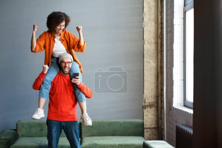 Photo for Delighted Caucasian bearded man holding mobile phone and his cute girlfriend on his shoulders, screaming and clenching their fists in excitement and happiness, winning the lottery, achieving goals - Royalty Free Image