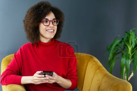 Téléchargez les photos : Belle femme ethnique portant des lunettes élégantes, assise sur un fauteuil dans un bureau moderne, utilisant le téléphone portable, textos, vérifier le contenu des médias sociaux, planifier des réunions, sourire en regardant de côté - en image libre de droit