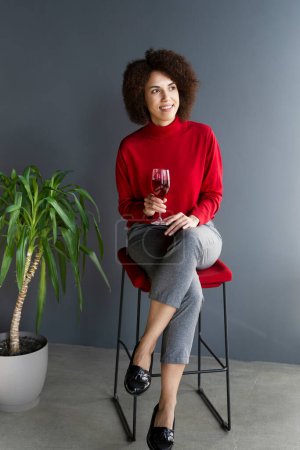 Foto de Retrato de cuerpo entero atractiva mujer multiétnica elegante y segura con pelo afro, sentado en una silla en un interior moderno, sosteniendo una copa de vino y tomando un sorbo de vino tinto, mirando pensativamente a un lado - Imagen libre de derechos