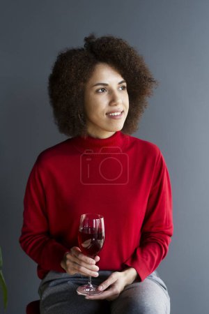 Foto de Retrato seguro de una hermosa mujer joven afroamericana elegante, vestida con suéter rojo, con una copa de vino, sentada en una silla, mirando cuidadosamente hacia otro lado, aislada sobre un fondo de pared gris - Imagen libre de derechos