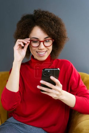 Foto de Mujer bonita afroamericana confiada con elegantes gafas enmarcadas rojas y suéter casual, comprobando el contenido de las redes sociales en su teléfono inteligente moderno, navegando por sitios web, sentada en un sillón cómodo - Imagen libre de derechos