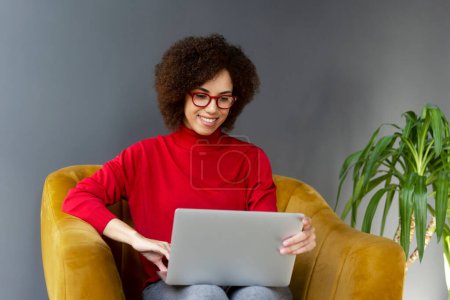 Foto de Joven mujer afroamericana gerente ejecutivo, mujer de negocios en línea de trabajo, escribiendo texto en el ordenador portátil, sentado en un sillón cómodo en una oficina corporativa contemporánea. Negocios. Gente. Tecnología - Imagen libre de derechos