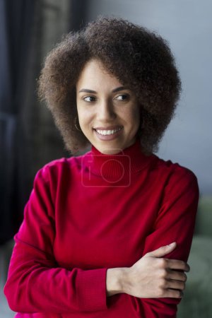 Photo for Stylish pensive African American young woman in red casual sweater, cutely smiling, looking to the side, standing with her arms folded in an office interior. Successful people. Business. Lifestyles - Royalty Free Image