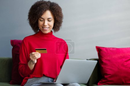 Foto de Sonriendo mujer afroamericana sosteniendo la tarjeta de crédito usando el ordenador portátil, compras en línea, buscando saldo de la tarjeta sentado en casa. Atractiva mujer pidiendo comida en el sitio web. Dinero electrónico, concepto de tecnología - Imagen libre de derechos
