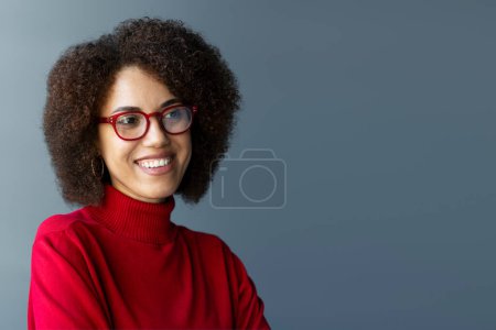 Foto de Retrato de una mujer de negocios afroamericana sonriente y confiada que lleva gafas con estilo y suéter de cuello alto rojo mirando hacia otro lado aislado en el fondo, espacio para copiar. Concepto de negocio exitoso - Imagen libre de derechos