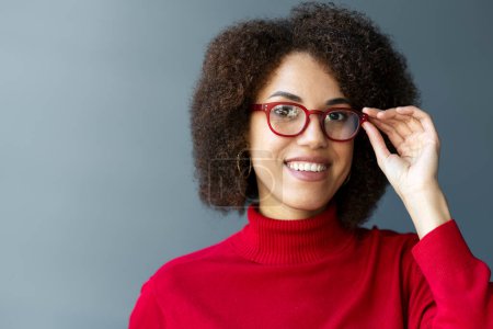 Foto de Sonriente mujer afroamericana con gafas con estilo y suéter de cuello alto rojo mirando a la cámara aislada en el fondo. Concepto de visión - Imagen libre de derechos