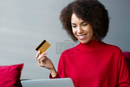 Foto de Sonriente mujer afroamericana sosteniendo la tarjeta de crédito usando el ordenador portátil de compras en línea, pidiendo comida en el sitio web sentado en casa. Concepto de venta - Imagen libre de derechos