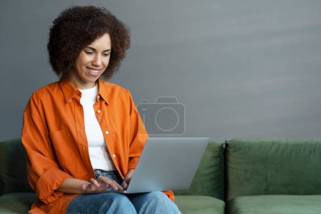 Téléchargez les photos : Sourire femme afro-américaine confiante en utilisant un ordinateur portable travaillant en ligne à partir de la maison. Rédacteur pigiste tapant sur le clavier, regardant des cours de formation, cherchant sur le site Web assis dans le bureau - en image libre de droit