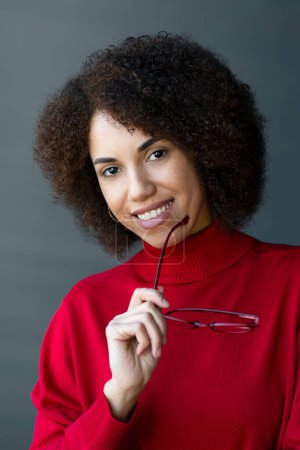 Photo for Portrait of attractive smiling African American woman holding red eyeglasses looking at camera isolated on background, vision concept. Fashion model in turtleneck sweater posing for pictures - Royalty Free Image