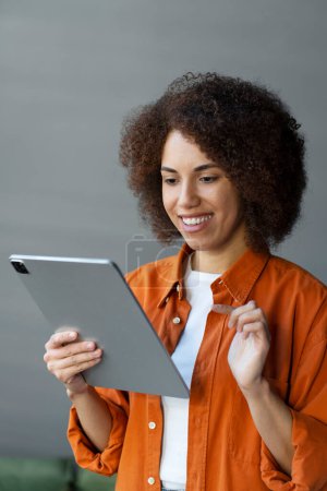 Téléchargez les photos : Souriant femme afro-américaine attrayante en utilisant des tablettes numériques achats en ligne, commander de la nourriture à la maison. Heureux succès confiant e livre de lecture féminine, regarder des vidéos en ligne. Concept technologique - en image libre de droit