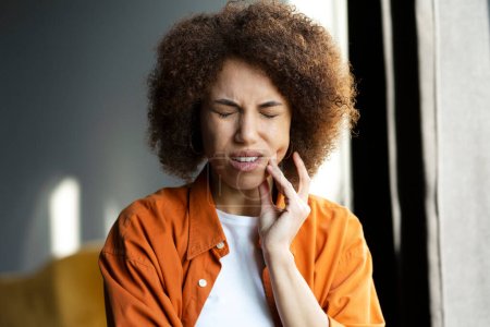 Téléchargez les photos : Triste femme afro-américaine malheureuse tenant la main sur sa joue ayant mal aux dents, besoin d'un traitement médical debout à la maison. Soins dentaires - en image libre de droit