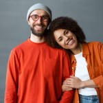 Happy stylish romantic couple embracing, looking at camera standing together at new home. Portrait of smiling attractive confident best friends wearing colorful clothing. Love, relationship concept