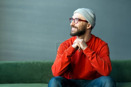 Foto de Hombre barbudo sonriente pensativo con gafas con estilo y sombrero hipster mirando hacia otro lado, la planificación de inicio sentado en el sofá en casa, espacio para copiar. Negocios exitosos - Imagen libre de derechos