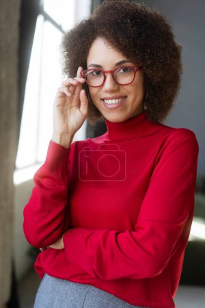 Foto de Atractiva mujer afroamericana sonriente con elegantes gafas rojas y suéter de cuello alto rojo mirando a la cámara aislada en el fondo. Retrato de estudiante inteligente y confiado. Concepto de visión - Imagen libre de derechos